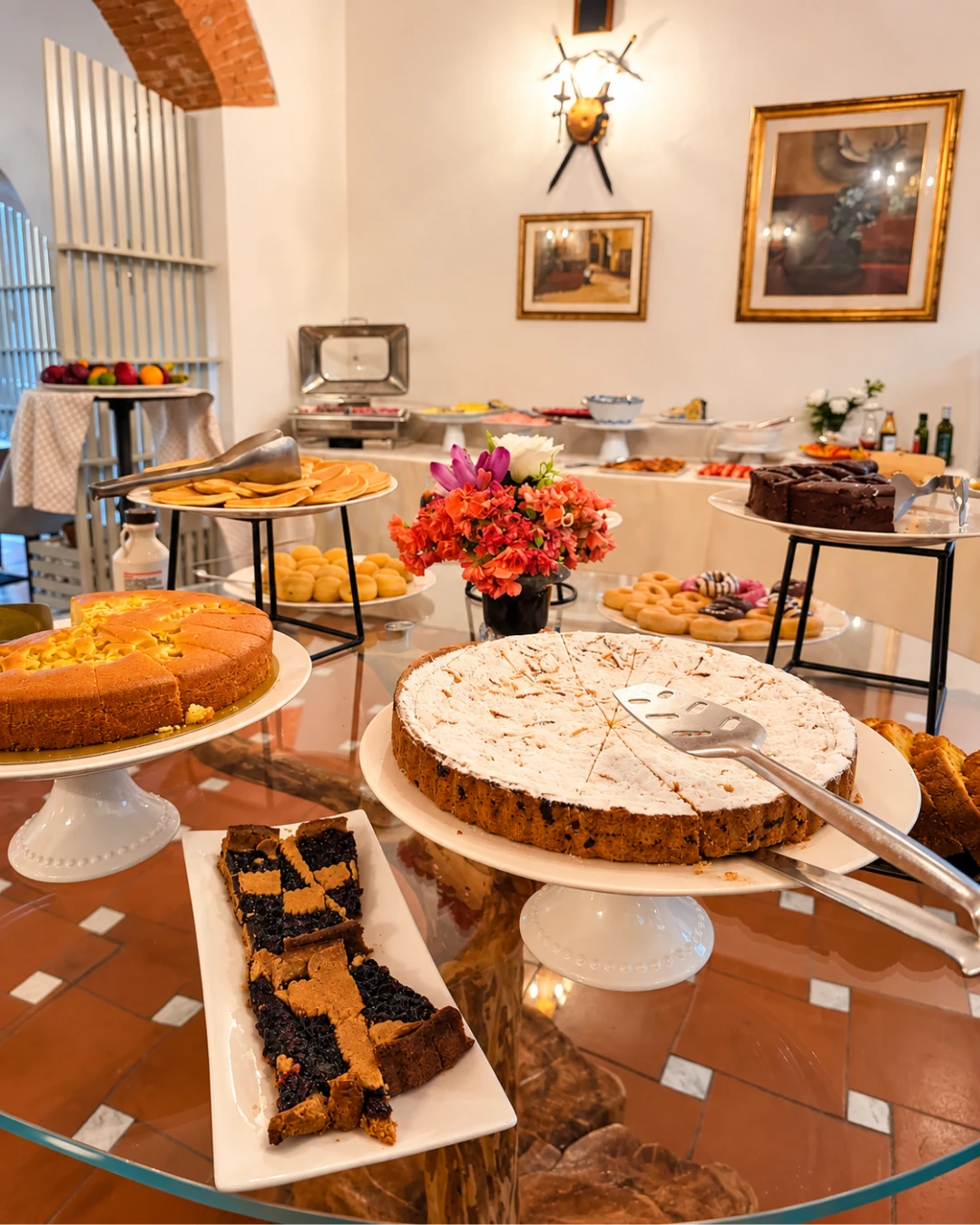 Il tavolo dei dolci: crostate, torte alle mandorle e un vaso di fiori, con il buffet salato sullo sfondo