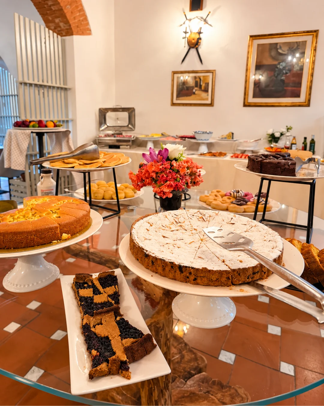 Il tavolo dei dolci: crostate, torte alle mandorle e un vaso di fiori, con il buffet salato sullo sfondo