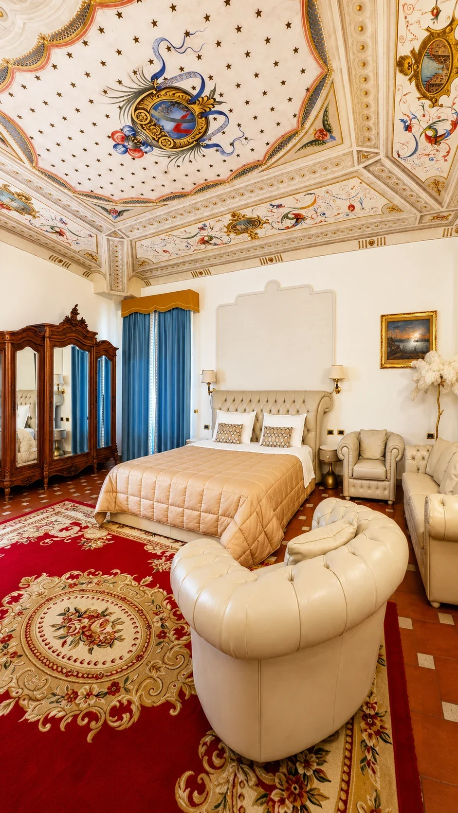 La Suite Ambrogi con letto capitonné, poltrone chiare, tappeto rosso e armadio in legno