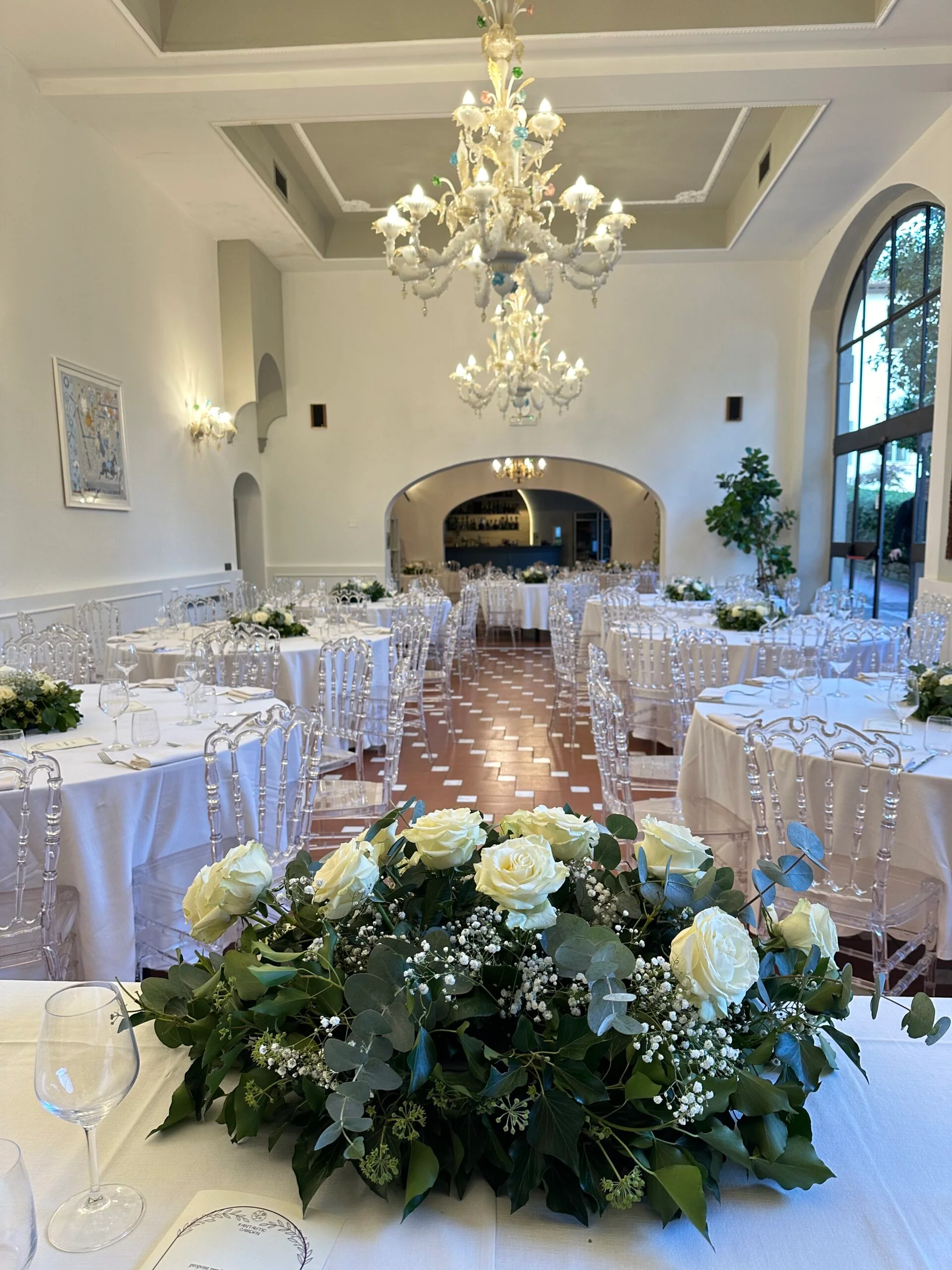 La sala della villa allestita per un matrimonio, con i tavoli bianchi e i centrotavola di rose sotto il lampadario di vetro