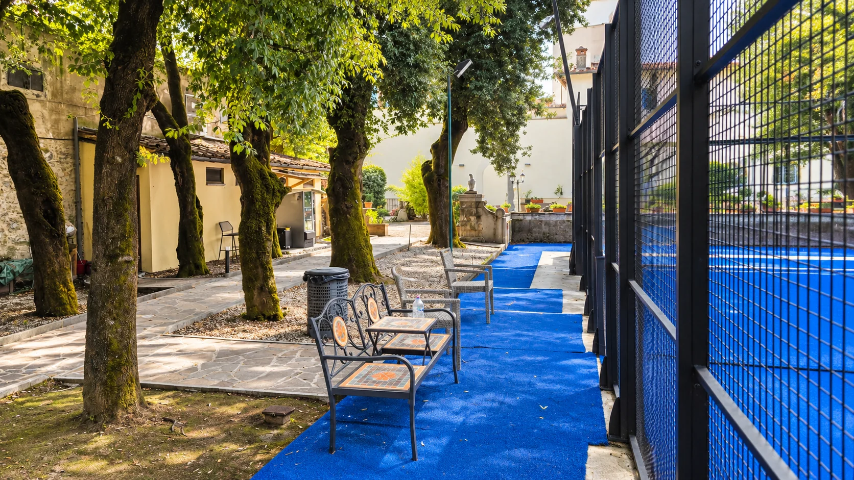Benches in the shade of the trees, along the blue walkway that runs beside the court