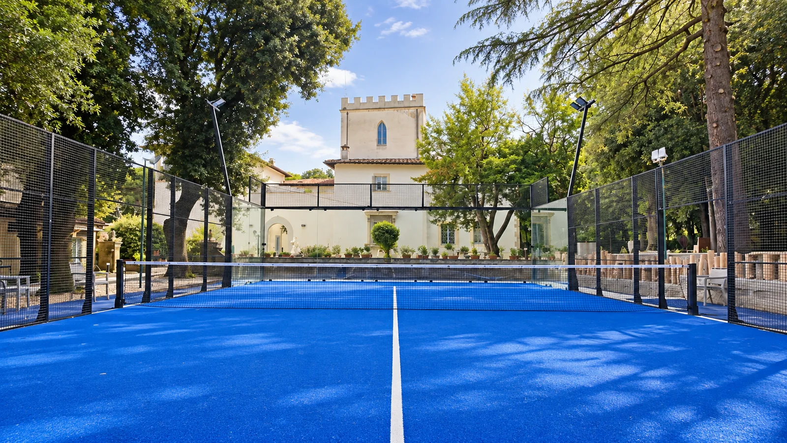 Il campo da padel visto dal fondo: il tappeto blu, la rete e, dietro le vetrate, la torre merlata della villa