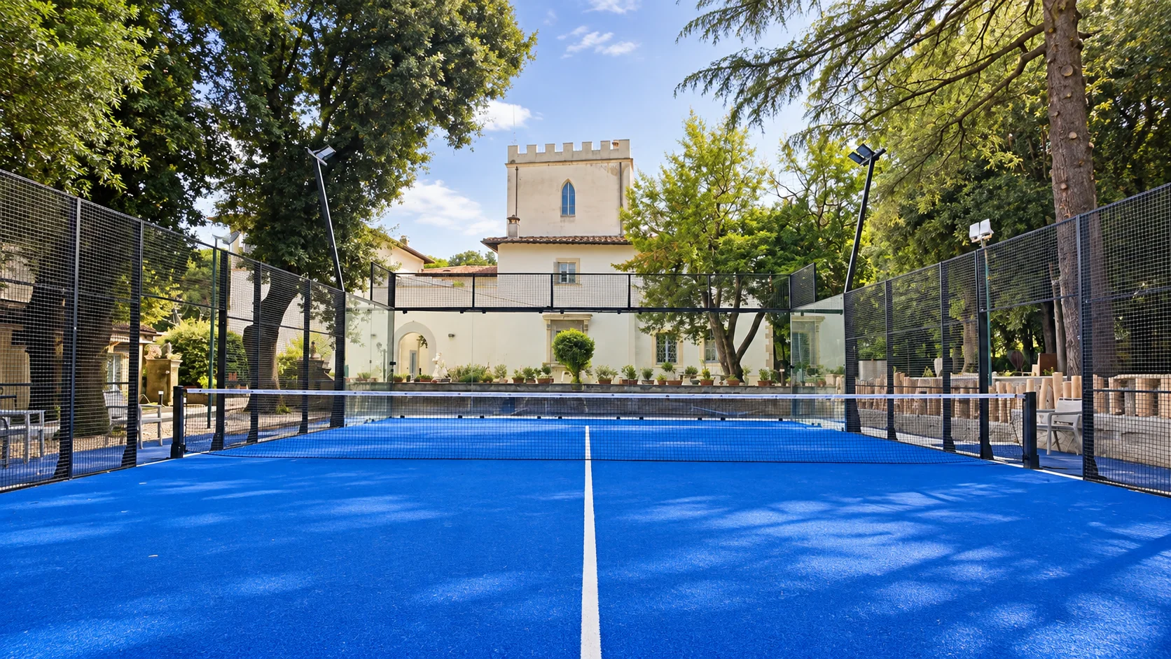 The padel court seen from the baseline: the blue surface, the net and, beyond the glass walls, the crenellated tower of the villa