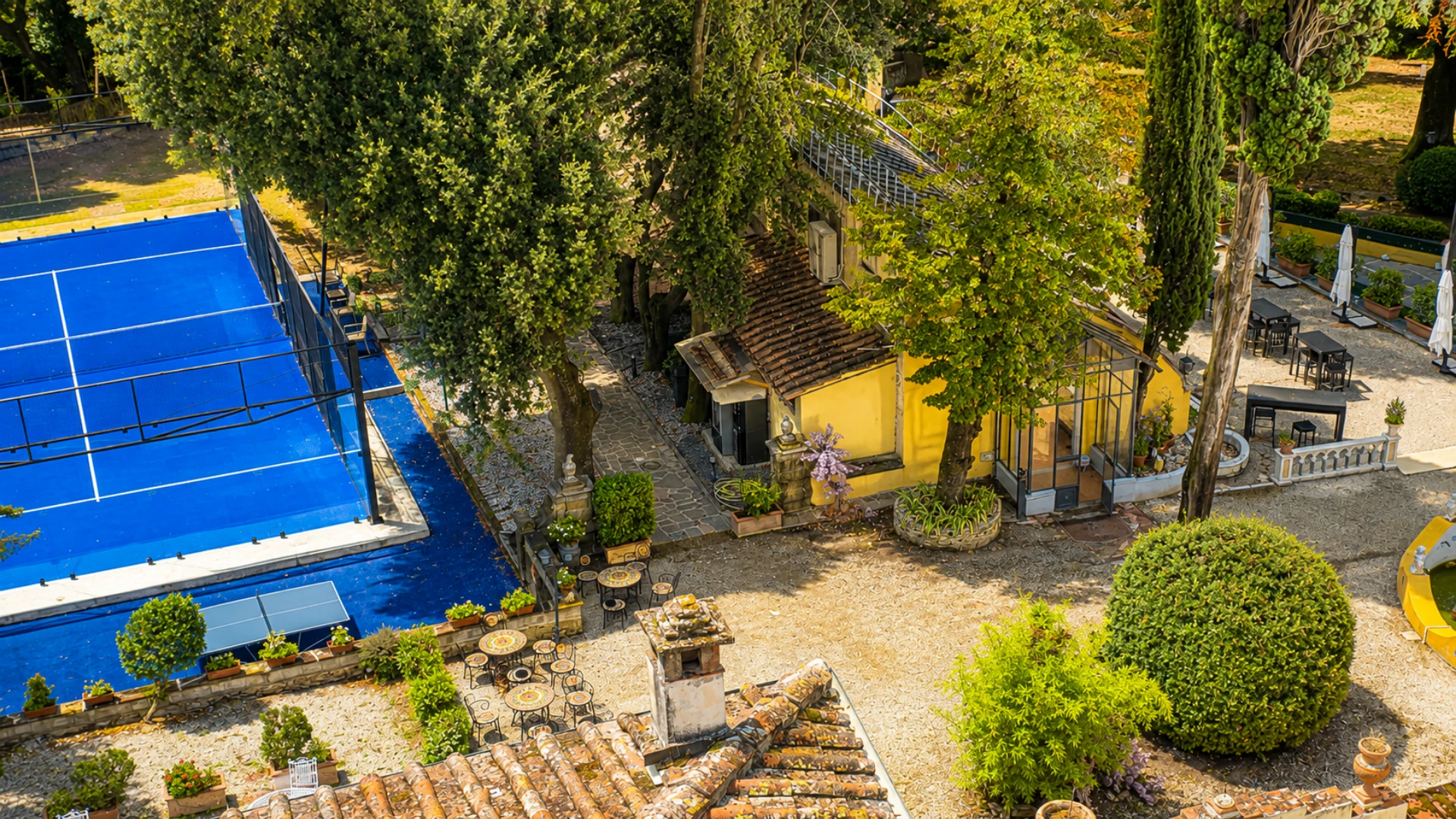 The padel court seen from above: its blue surface among the trees of the park, tables in the shade and the villa's yellow limonaia beside it