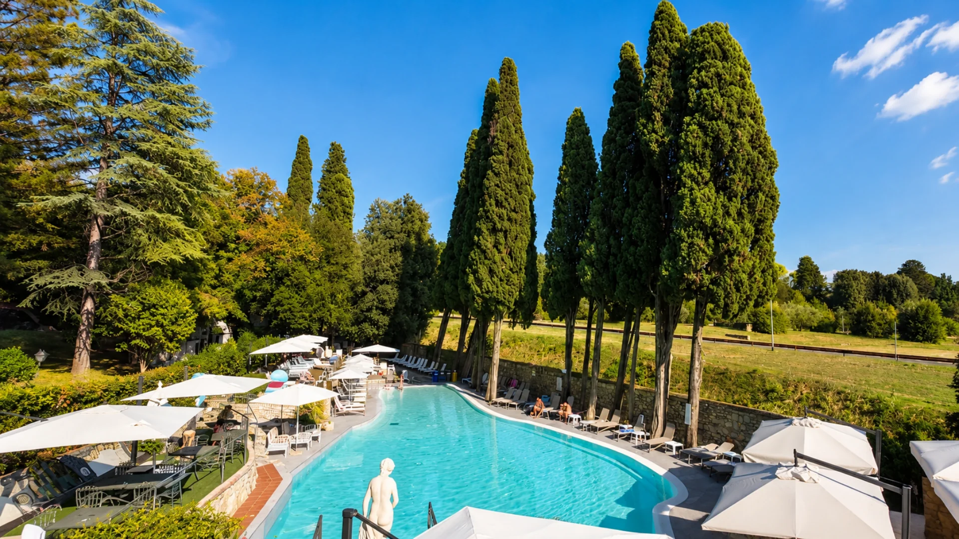 La piscina vista dall'alto: la vasca azzurra fra i cipressi, gli ombrelloni bianchi lungo il bordo, i tavolini all'ombra e una statua in pietra in primo piano