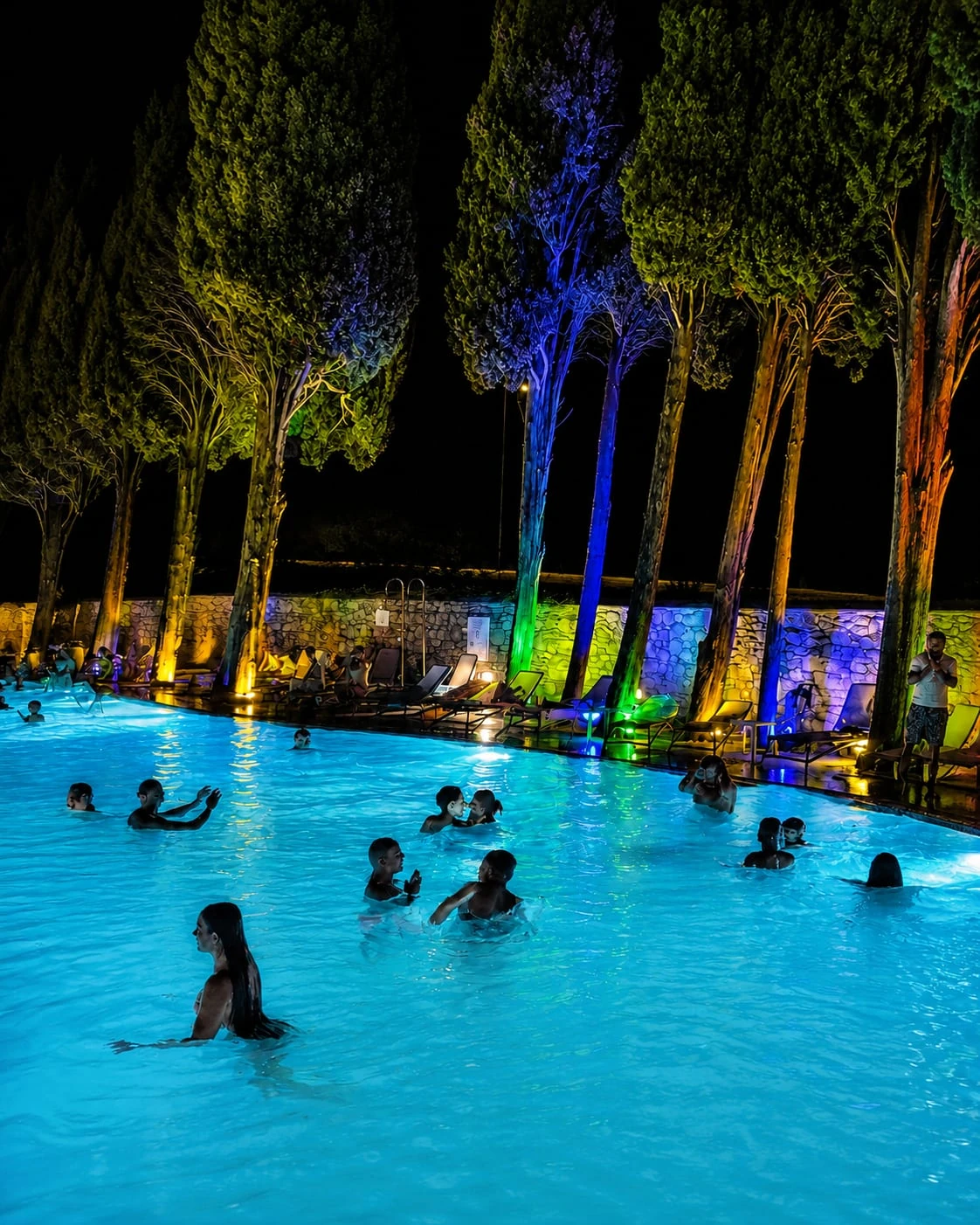 La piscina di notte, con i cipressi illuminati di blu, verde e arancio