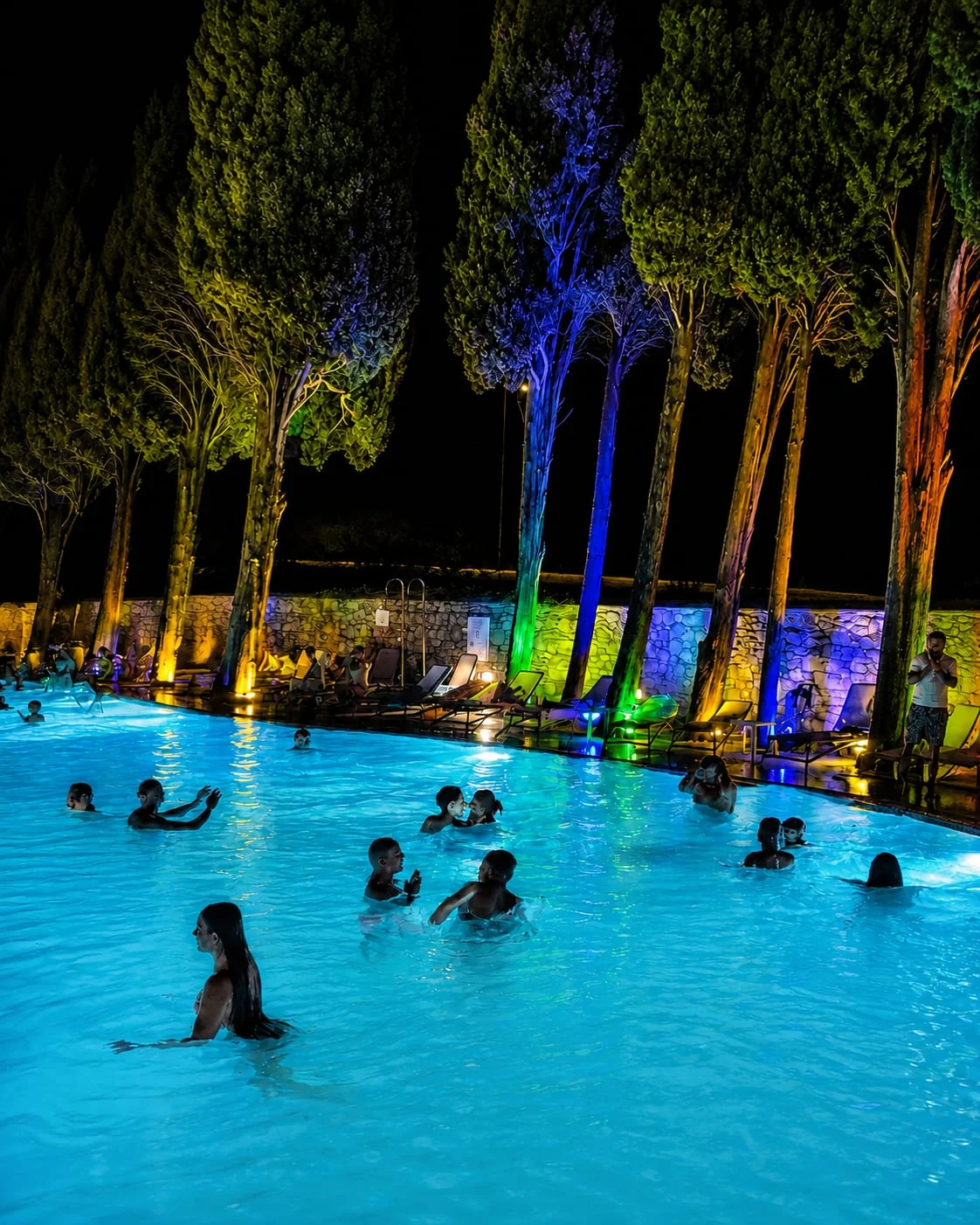 La piscina di notte, con i cipressi illuminati di blu, verde e arancio