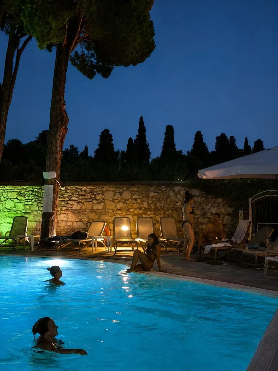 The pool lit up at night, with the stone wall and loungers along the edge