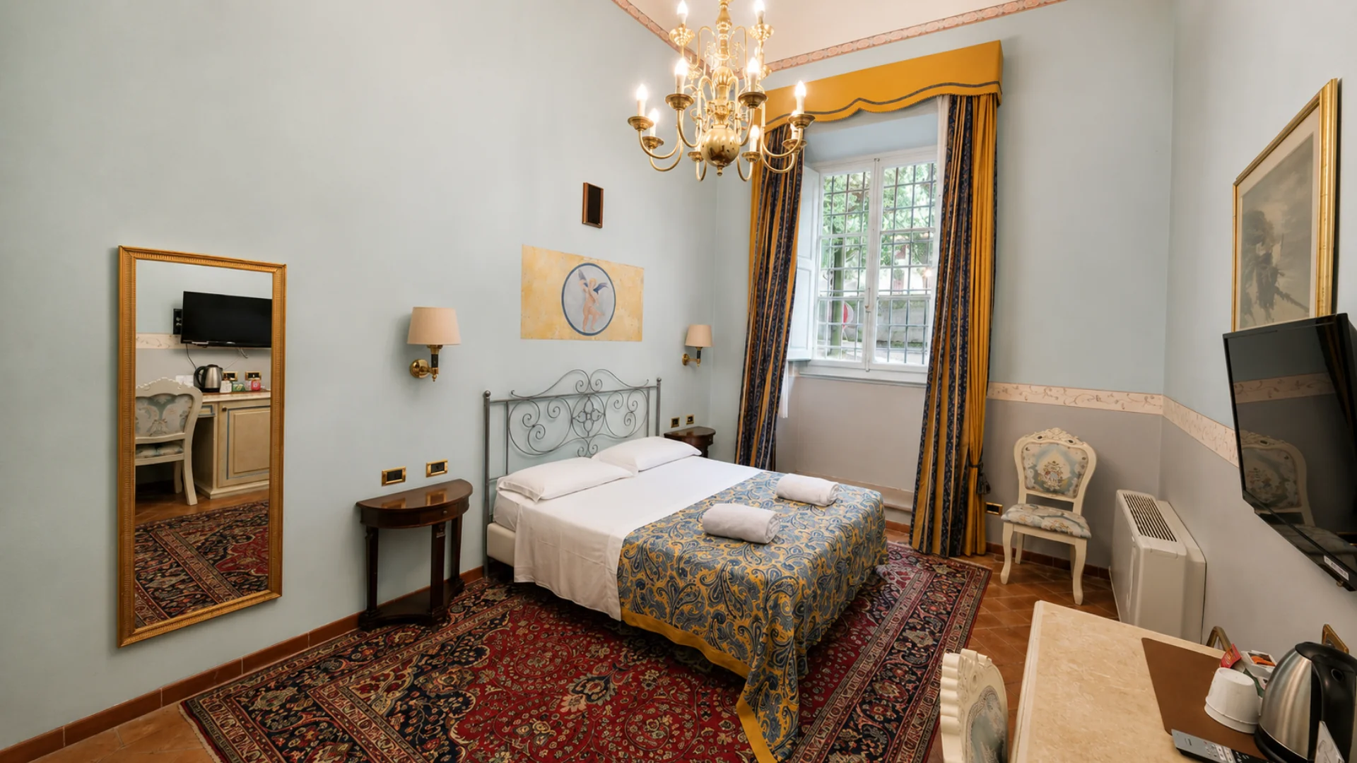 A pale blue Premium room with a wrought iron bed, a branched chandelier and yellow and blue curtains