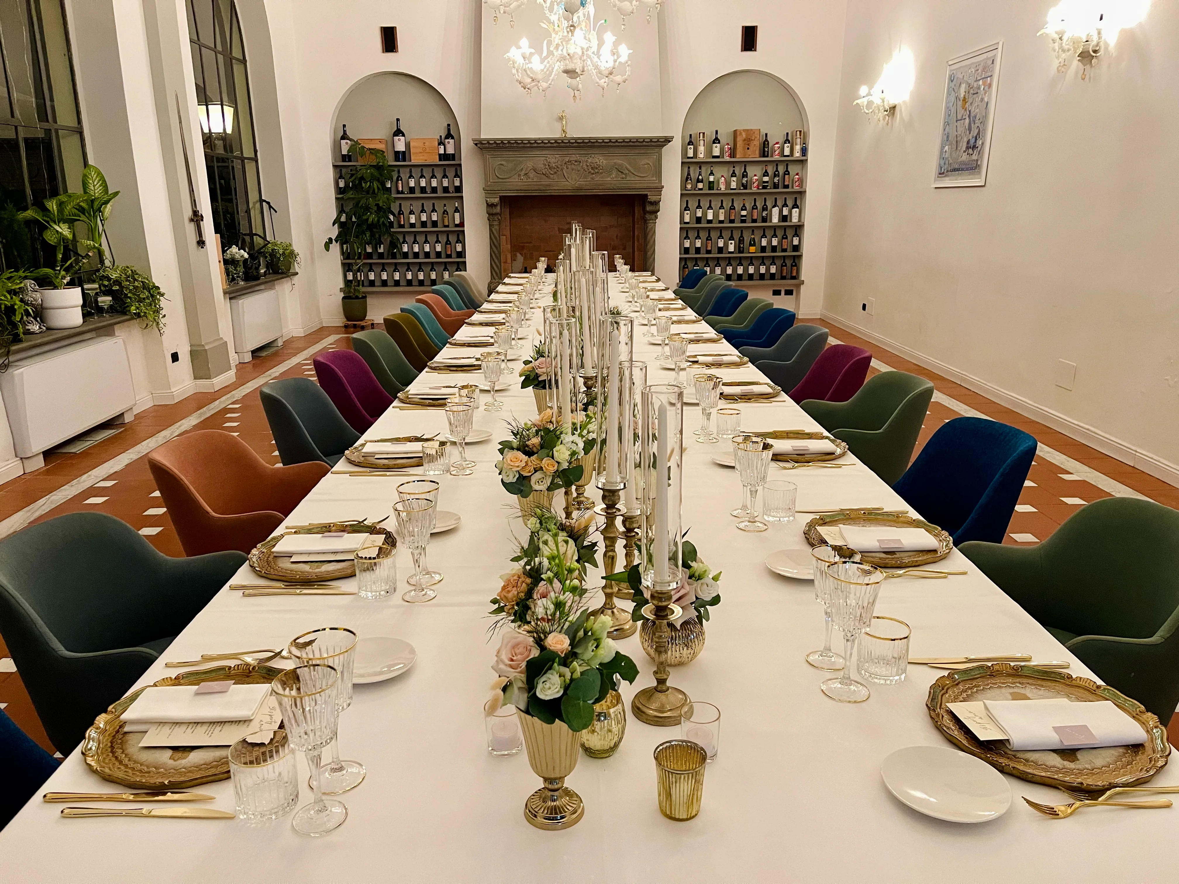 La sala del ristorante apparecchiata per un banchetto: la tavolata lunga con i candelabri e i fiori, le poltroncine di velluto colorato, il camino e gli scaffali dei vini