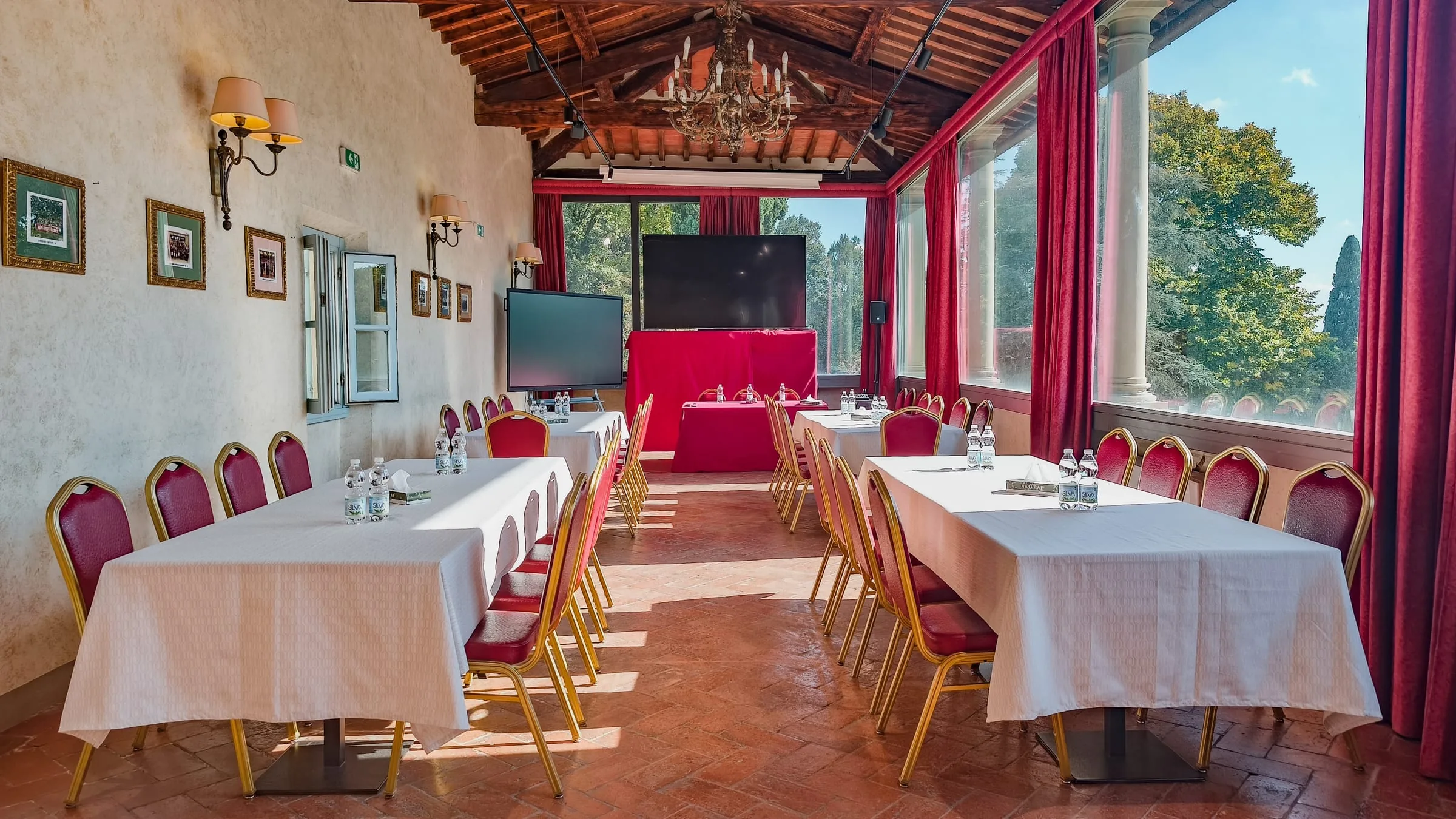 La sala meeting allestita a ferro di cavallo sotto la volta di travi: tavoli con le tovaglie chiare, sedie rosse e oro, il grande schermo in fondo e le vetrate con le tende rosse sul parco