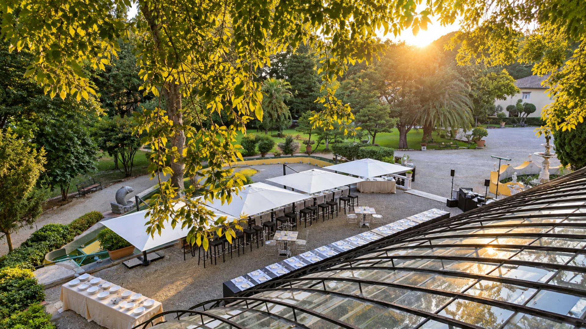 La terrazza vista dall'alto in controluce fra i rami: gli ombrelloni bianchi, i tavoli apparecchiati e il parco con la villa dietro