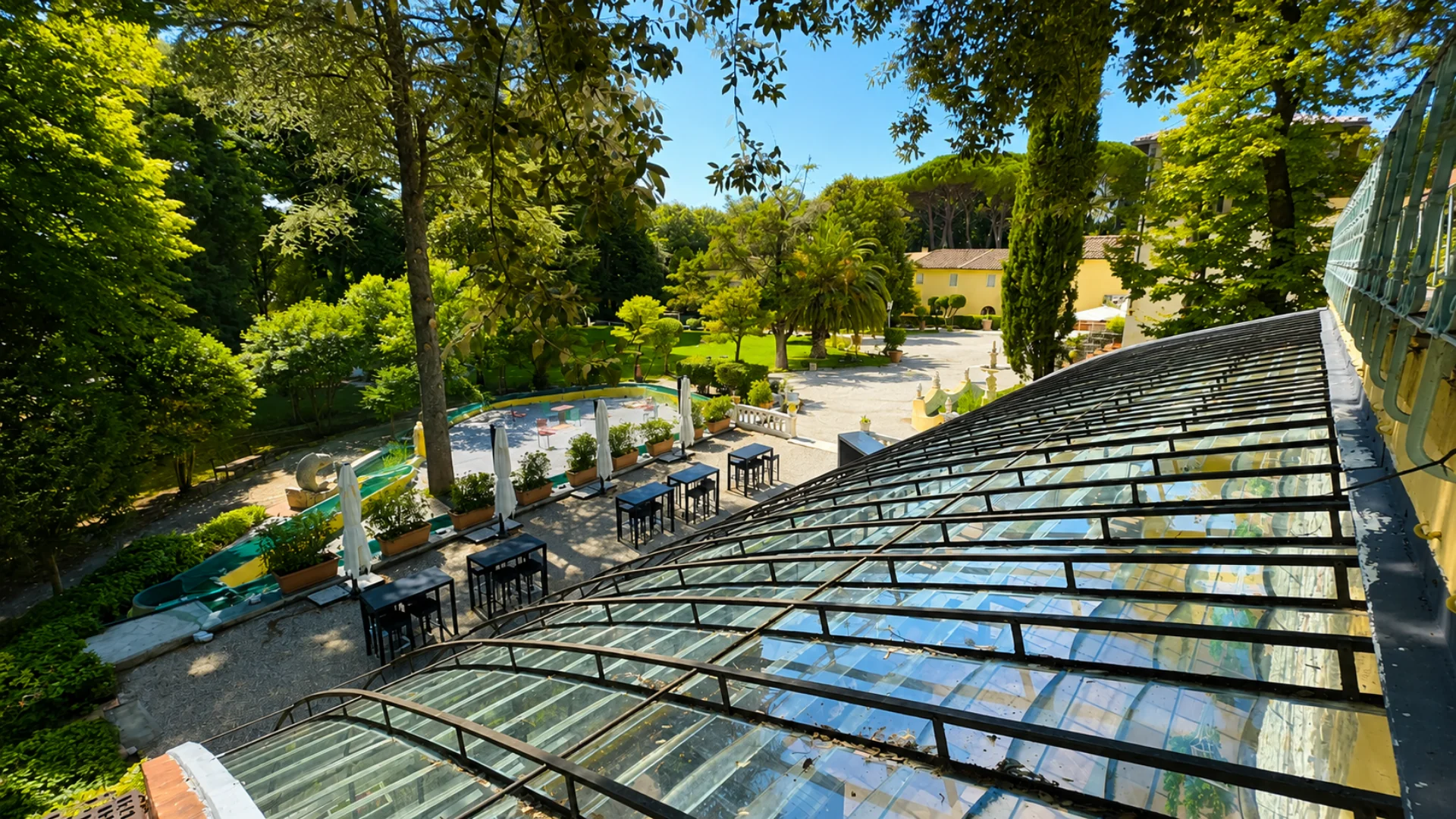 La volta a vetri vista dall'alto, e sotto la terrazza in ghiaia con i tavoli alti e gli ombrelloni chiusi, il parco e la villa sullo sfondo