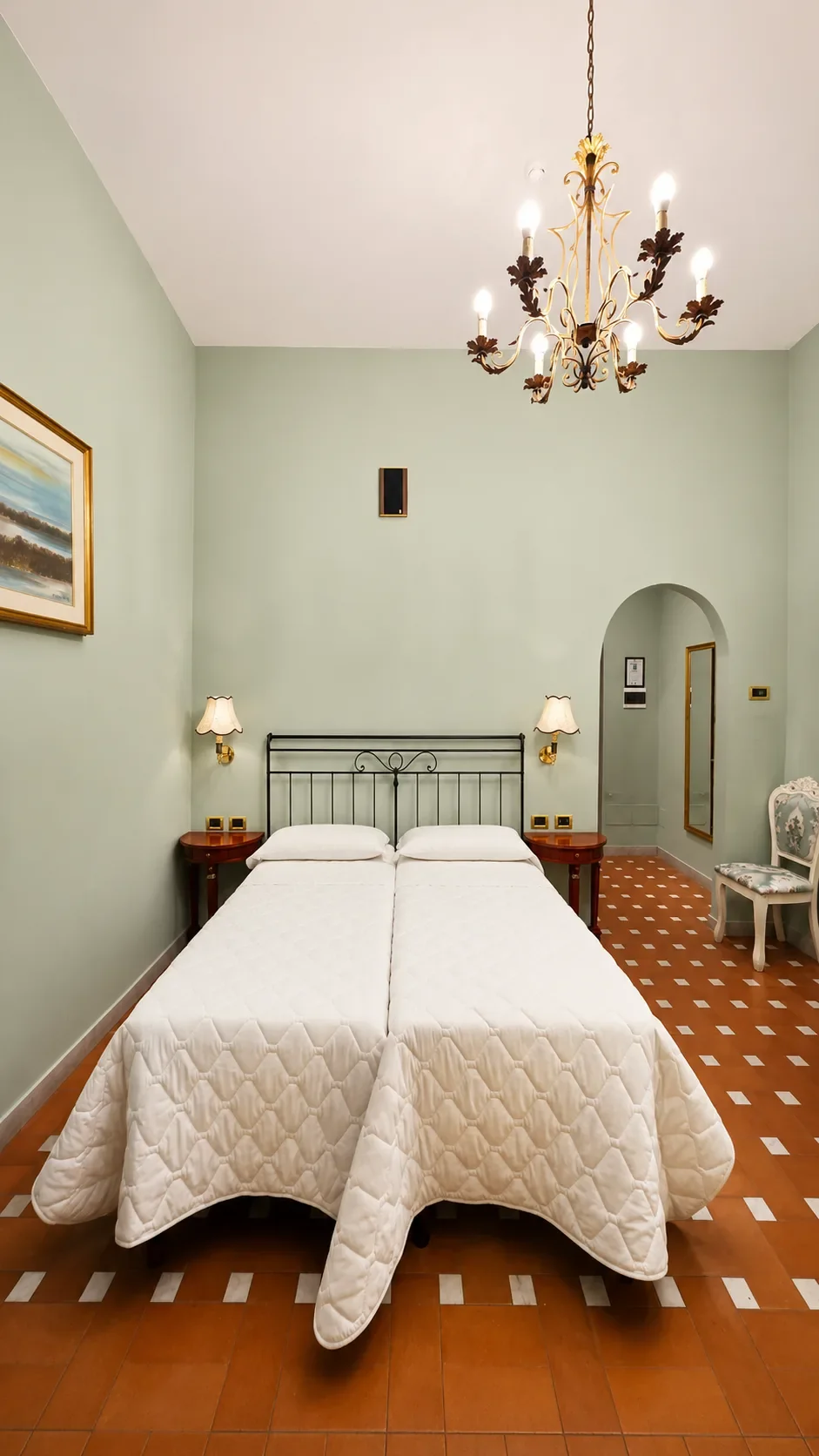 I due letti singoli della Camera Aletheia sotto il lampadario dorato, tra comodini in legno e pareti verde pastello